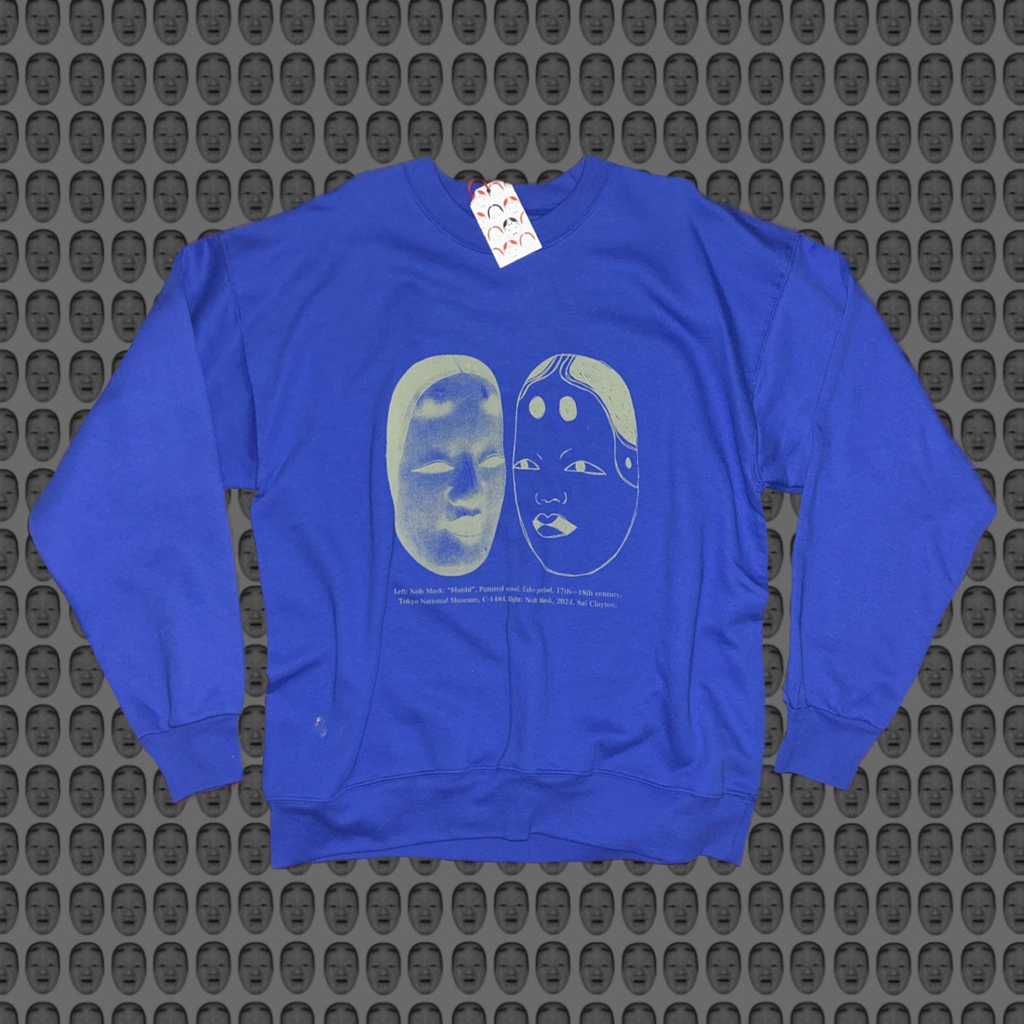 Blue sweatshirt with a matcha green face and noh mask design by STUDIO SAISAI on the front against a patterned background