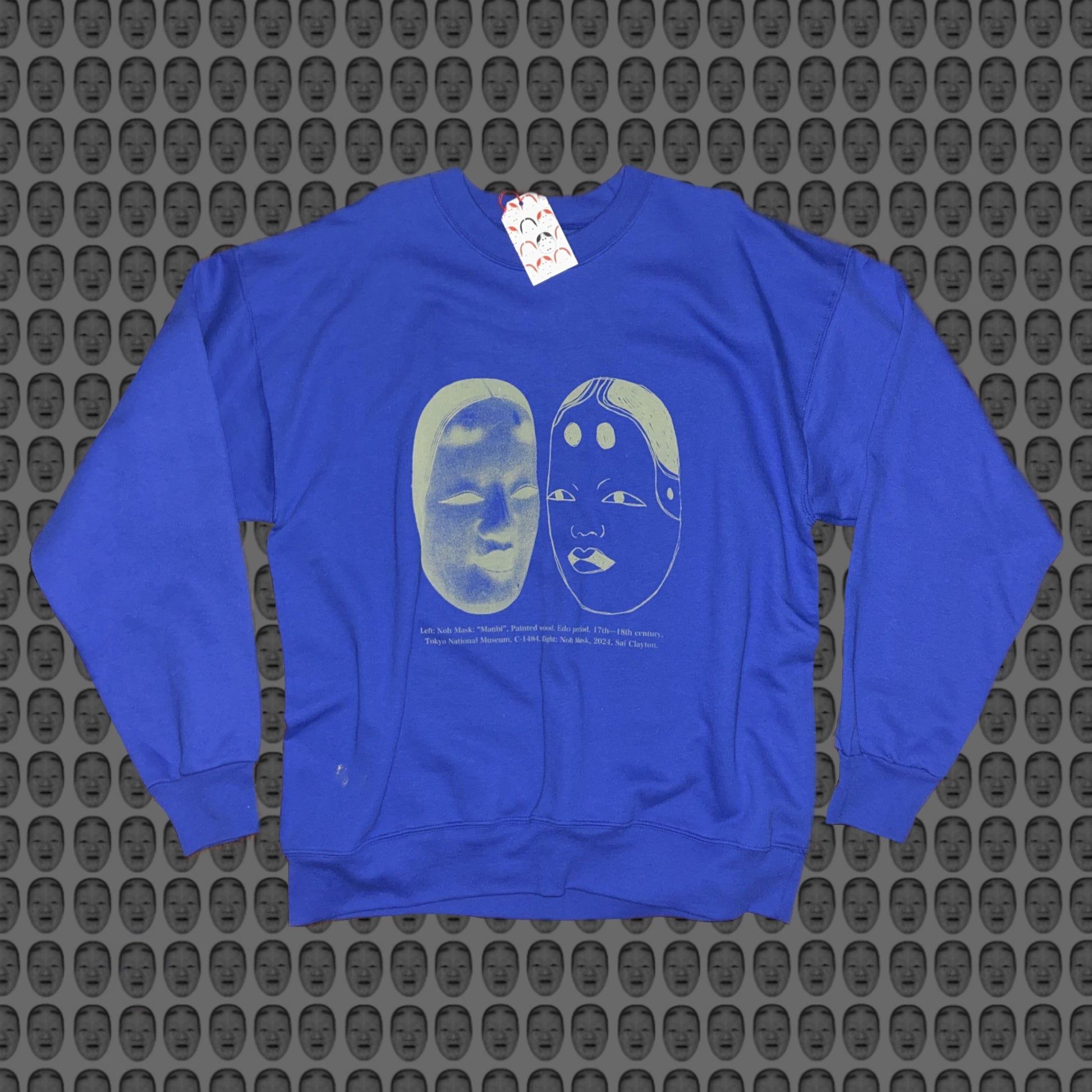 Blue sweatshirt with a matcha green face and noh mask design by STUDIO SAISAI on the front against a patterned background
