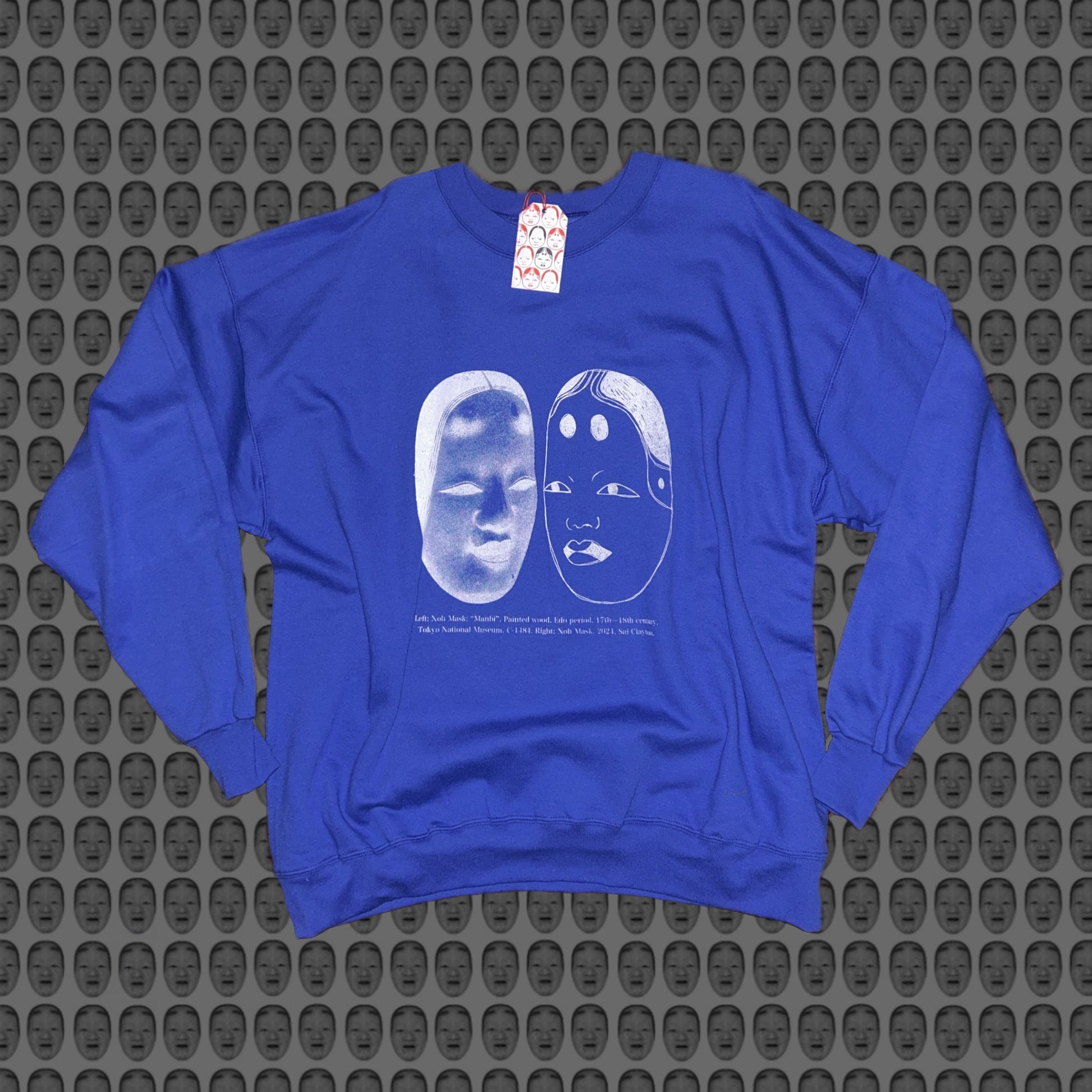 Blue sweatshirt with a white face and noh mask design by STUDIO SAISAI on the front against a patterned background