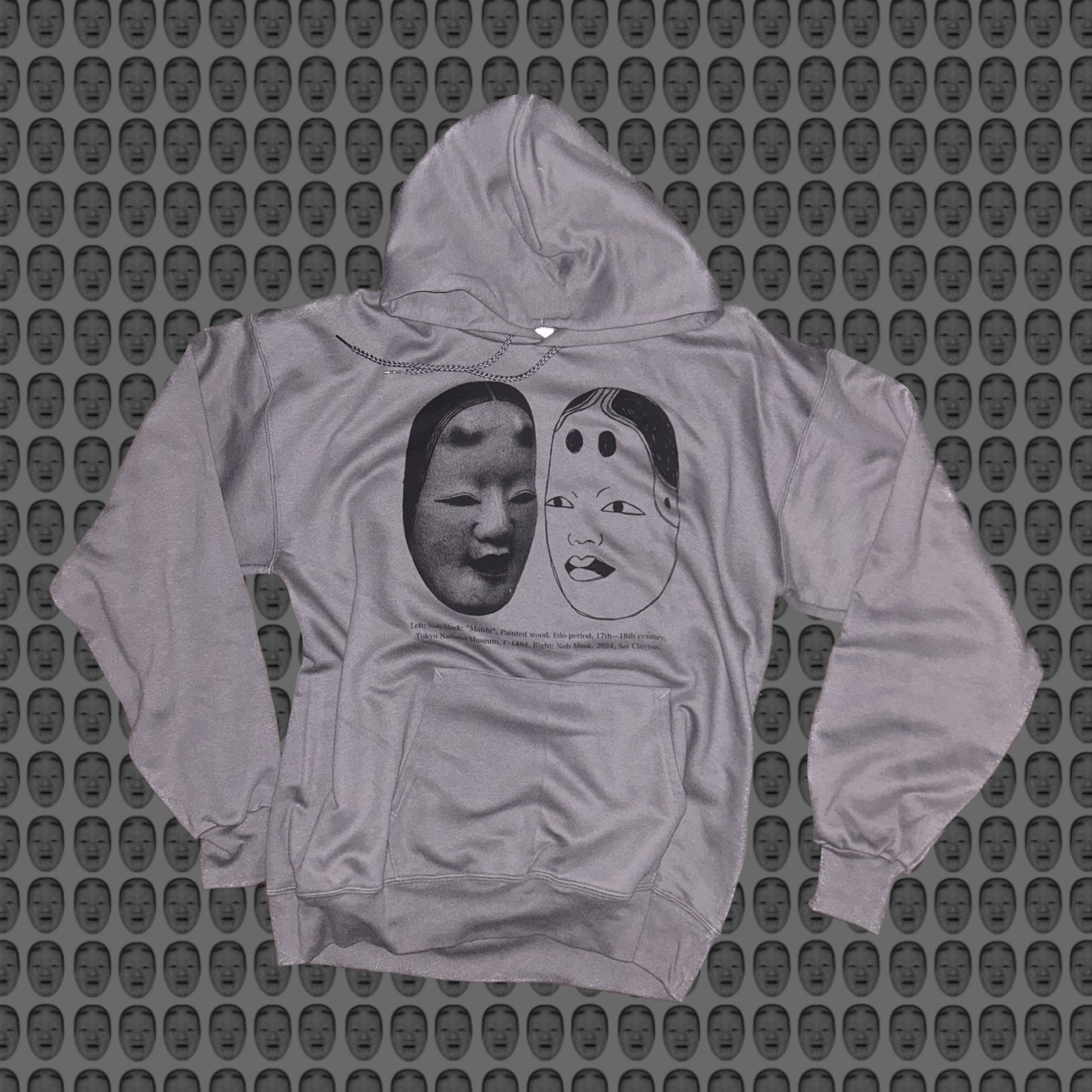 Gray hoodie with a face and noh mask design by STUDIO SAISAI on the front against a patterned background