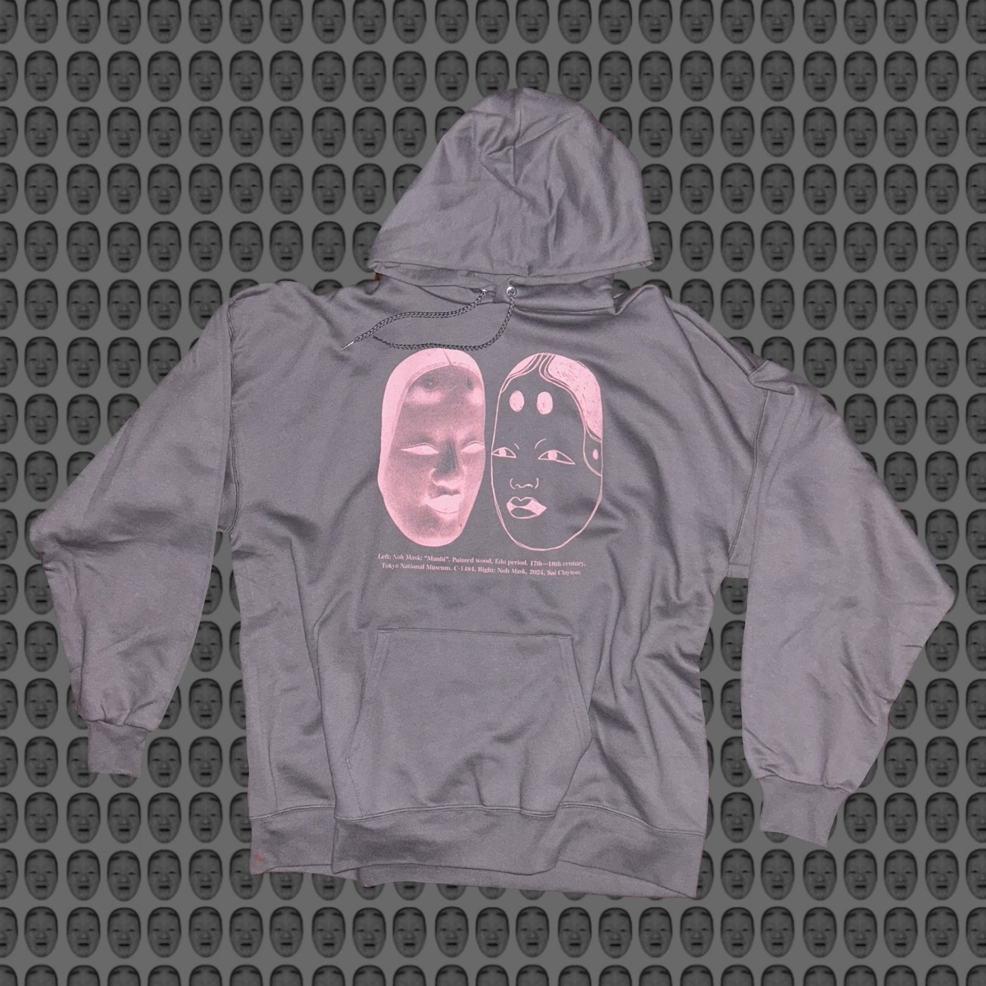 Gray hoodie with a pink face and noh mask design by STUDIO SAISAI on the front against a patterned background