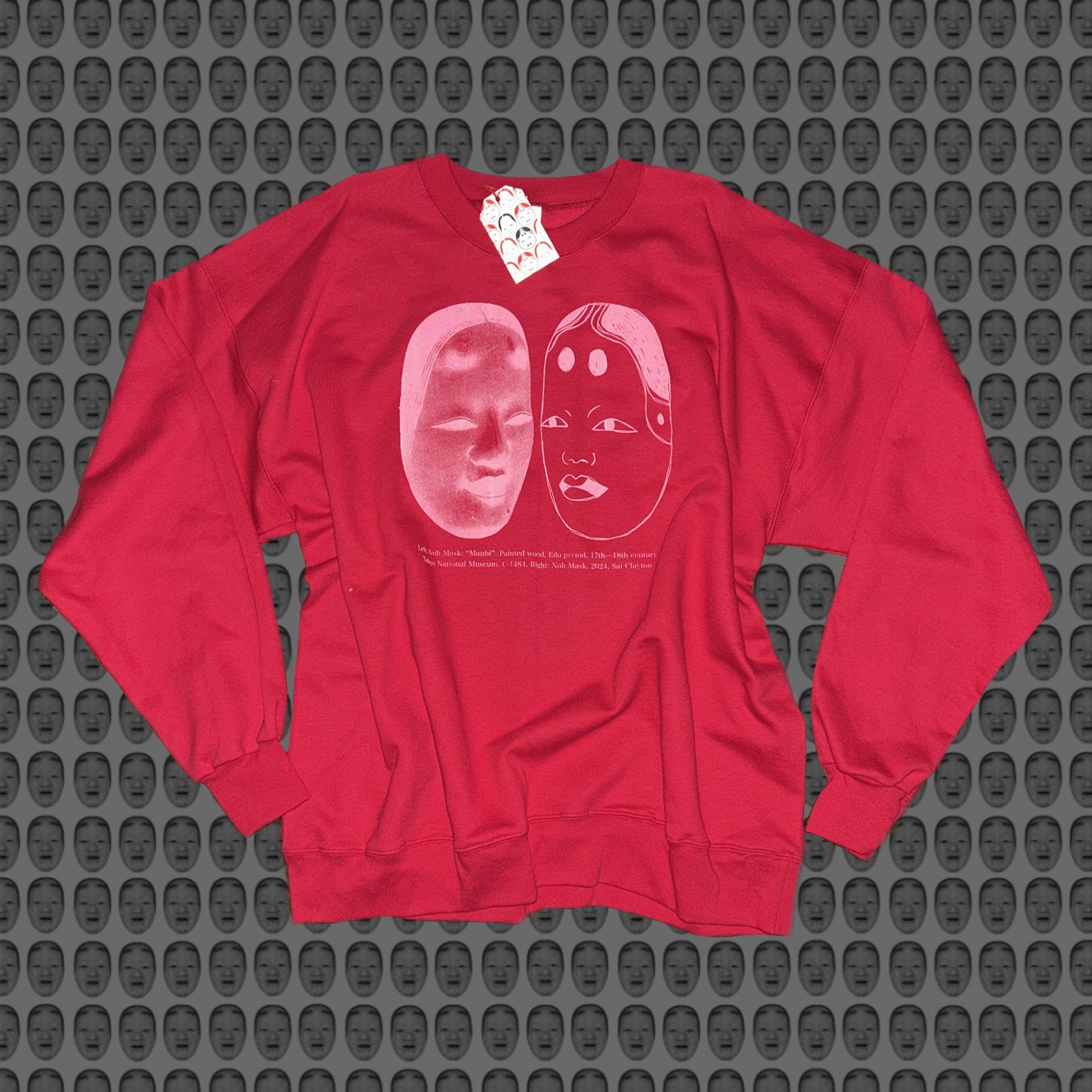 Red sweatshirt with a pink face and noh mask design by STUDIO SAISAI on the front against a patterned background